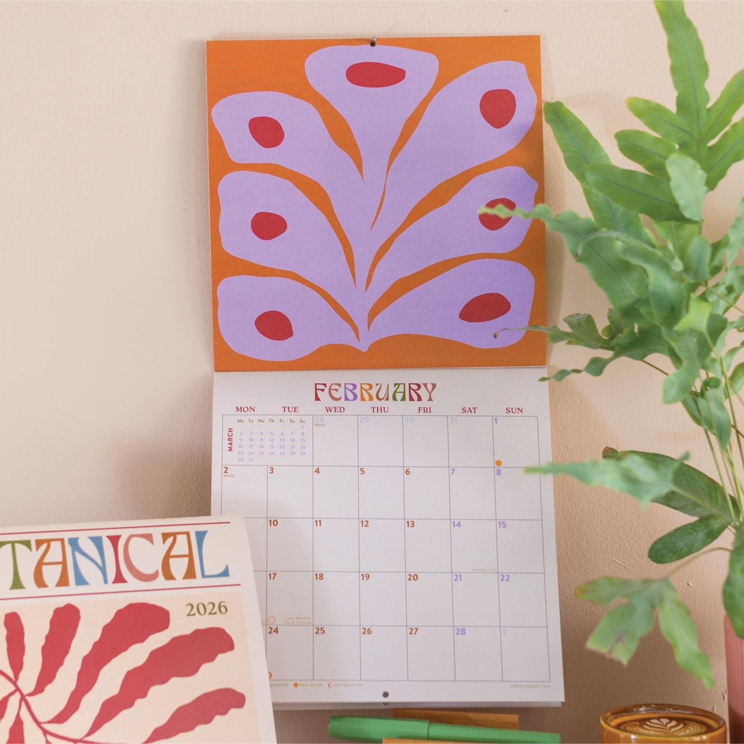 Calendar open to Febraury with a floral design and a plant on a light-colored wall