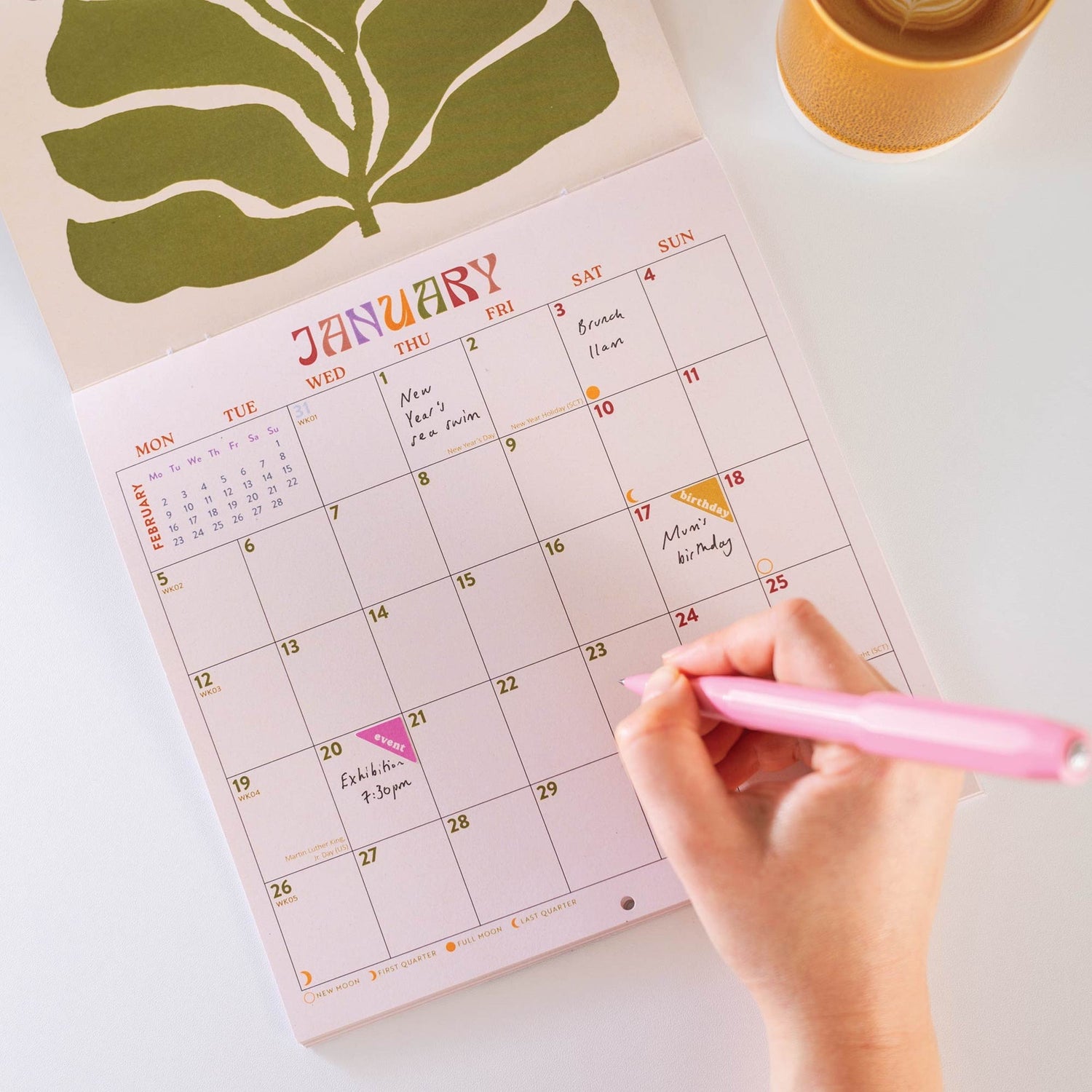 Hand holding a pink pen pointing to a date on a January calendar with a modern botanical illustration on the cover
