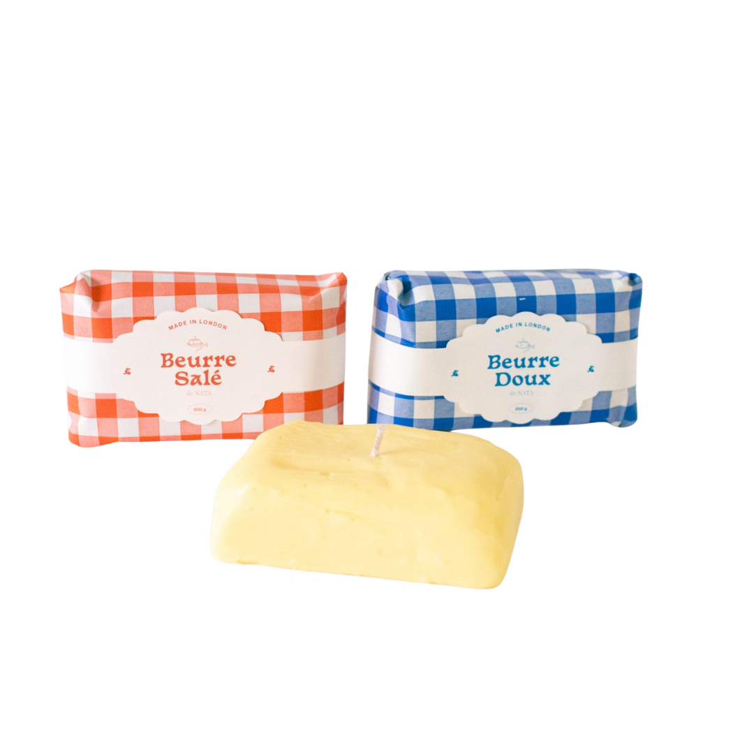 Two butter-style candles, one labeled Beurre Salé (salted) and the other Beurre Doux (unsalted), wrapped in blue and red gingham greaseproof paper and displayed on a white background.