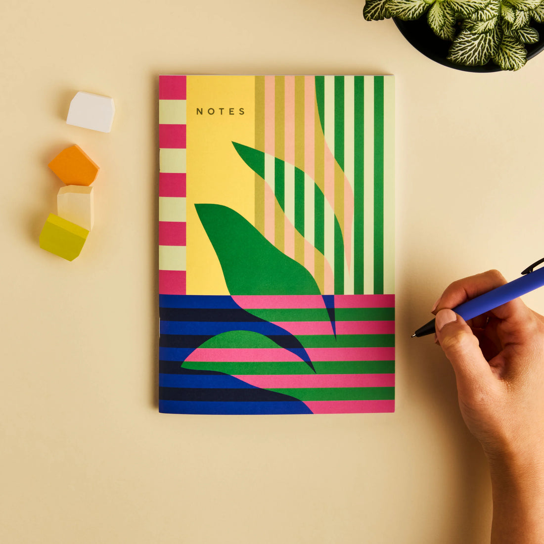 Colorful notebook with geometric design on a beige surface, hand holding a pen.
