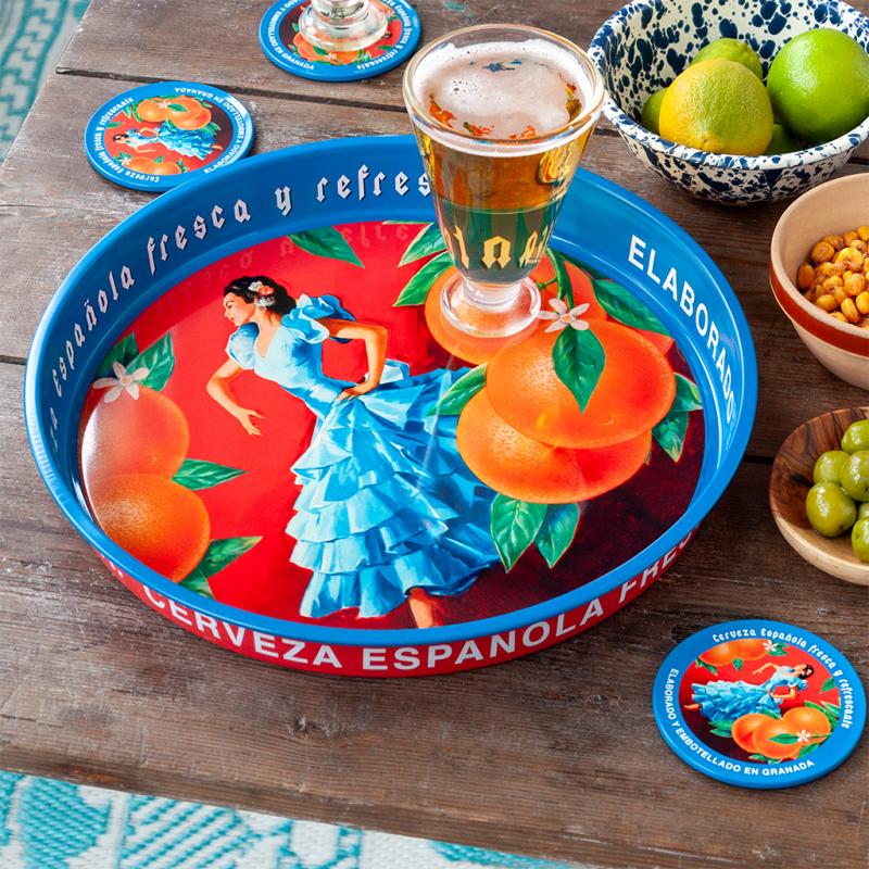 Decorative tray with Spanish-themed design featuring a woman in a blue dress and oranges, accompanied by a glass of beer on a wooden table.