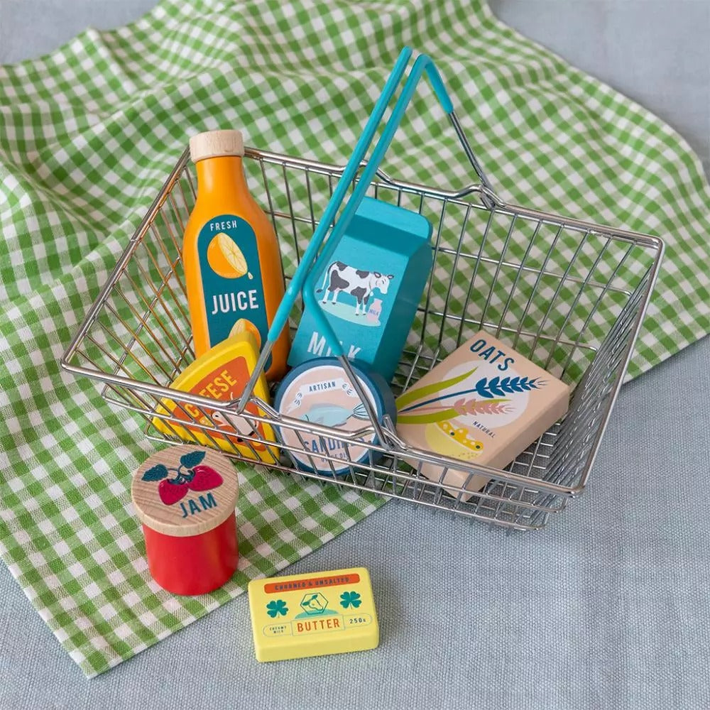 A metal shopping basket with rubberised plastic handles, containing various wooden pretend grocery items like milk carton, triangle cheese, oats, orange juice, and others.