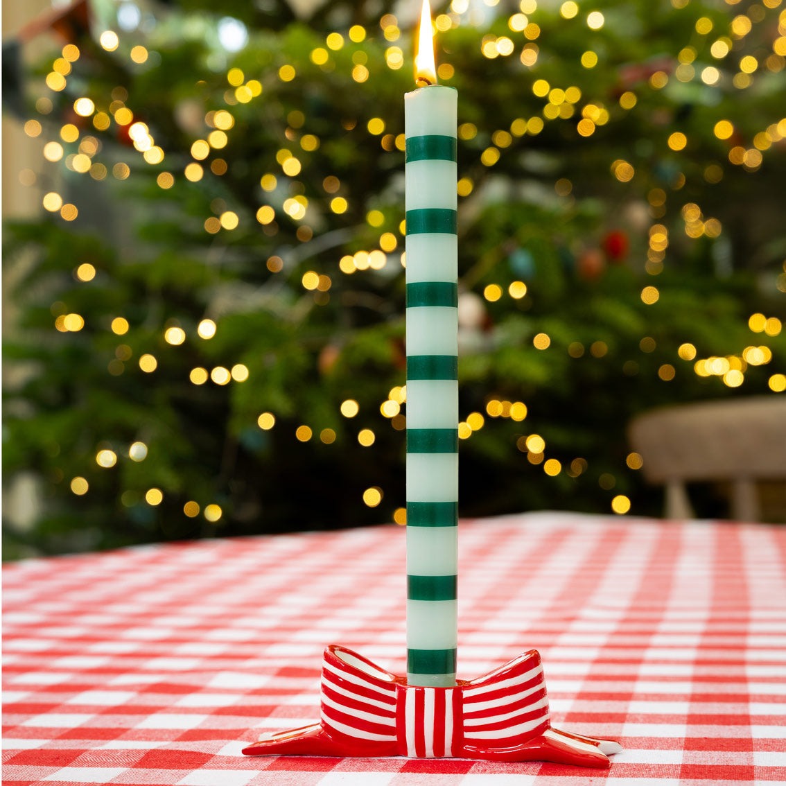 Green and white striped candle on a red and white striped bow candle holder with a blurred Christmas tree in the background.
