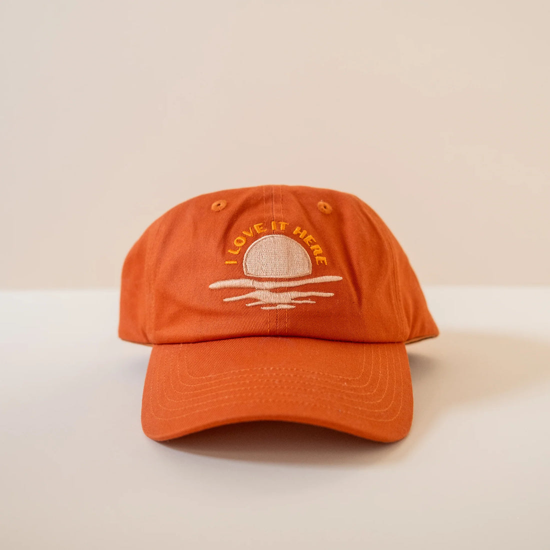 Burnt orange baseball cap with embroidered text "I love it here" and sun and wave design on the front