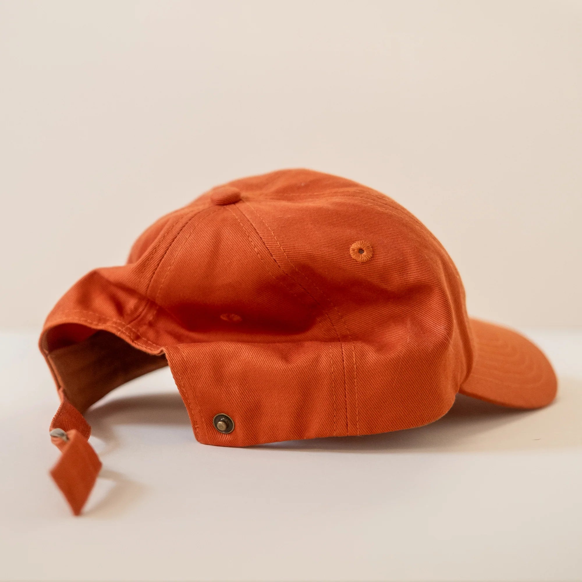 Orange cap with embroidered design on the front, featuring an adjustable brass closure and a back popper button for ponytail access.