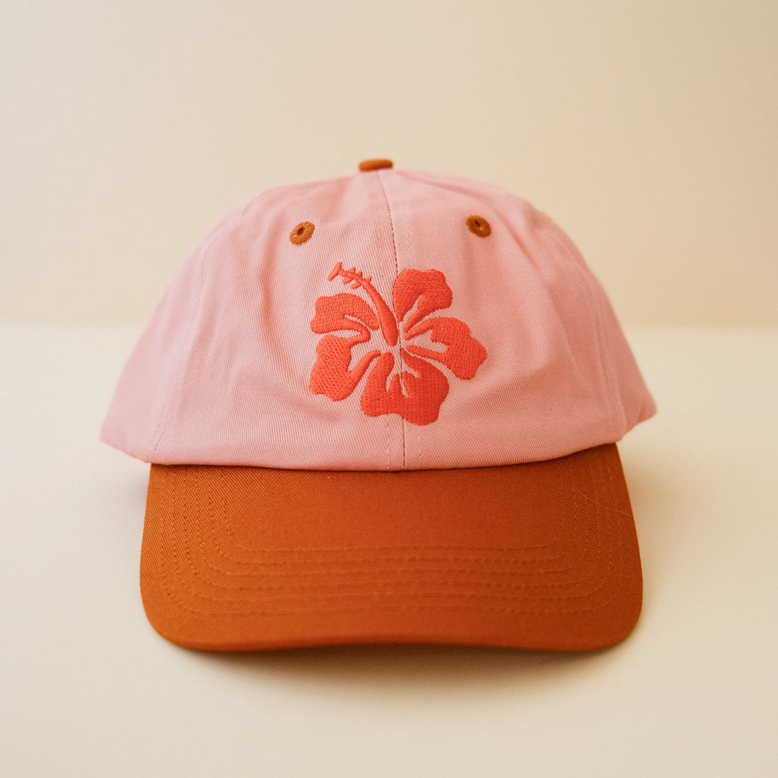 A pink cap with a burnt orange hibiscus embroidery on the front on a beige background