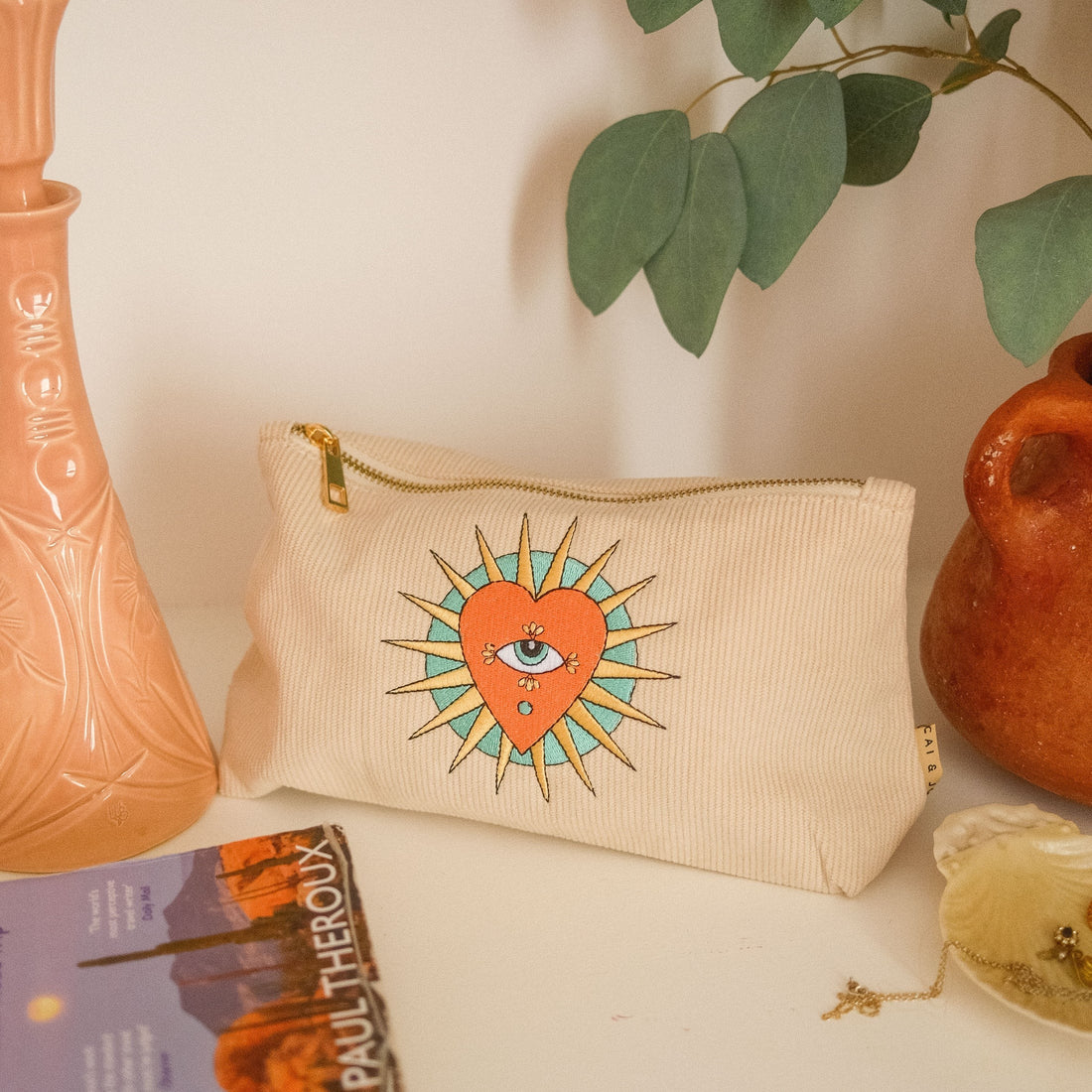 A beige corduroy pouch with a decorative pattern featuring a sun, heart and an eye on the front.