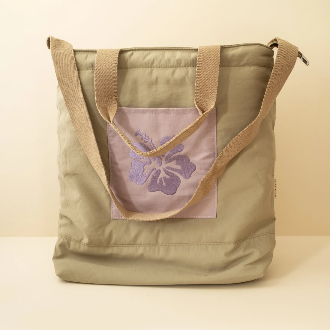 Olive tote bag with two brown handles and a purple embrodery flower design on the front pocket on a beige background