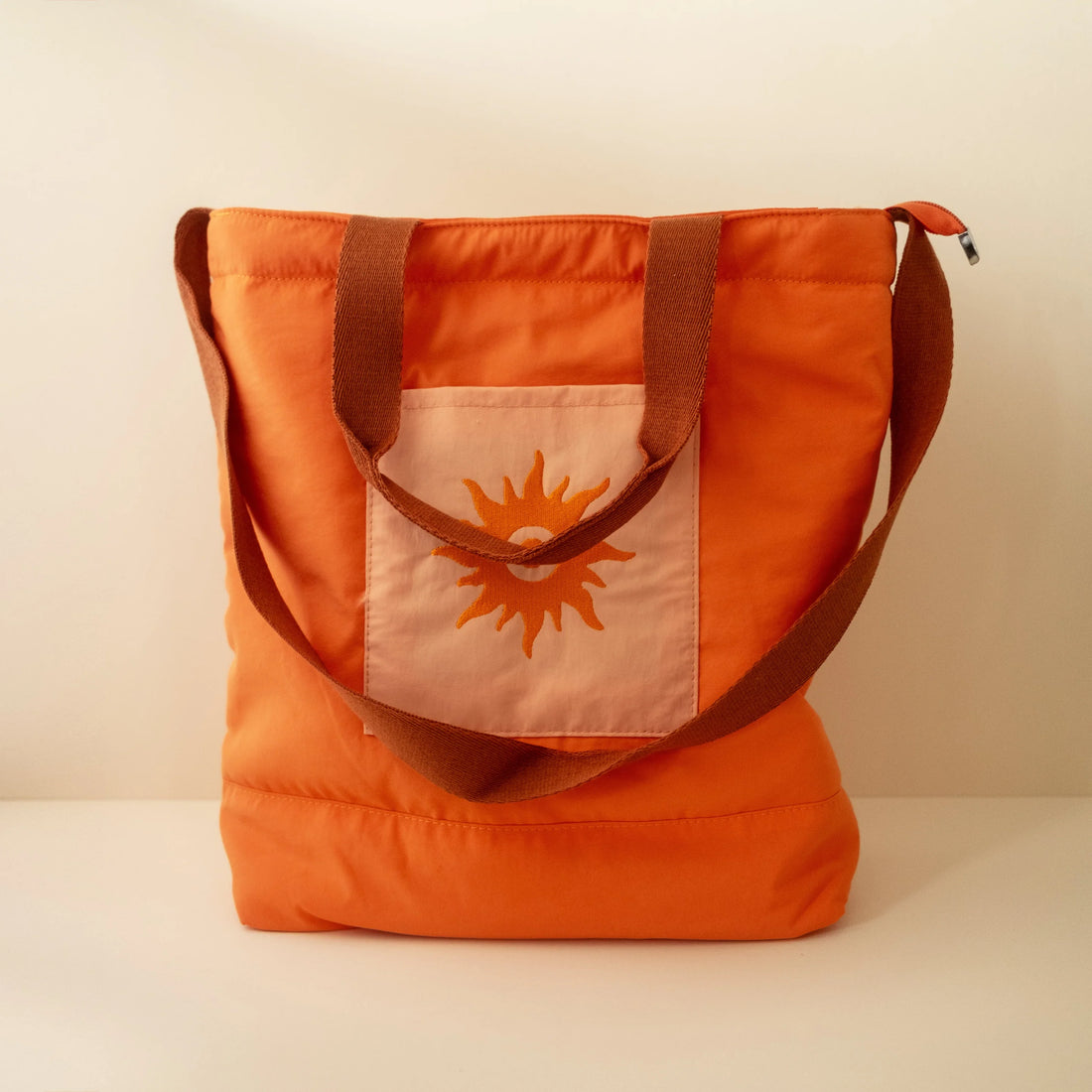 A burnt orange nylon beach tote with a sun pattern, featuring a top handle, a zipper closure, and an embroidered pocket on the front on a beige background