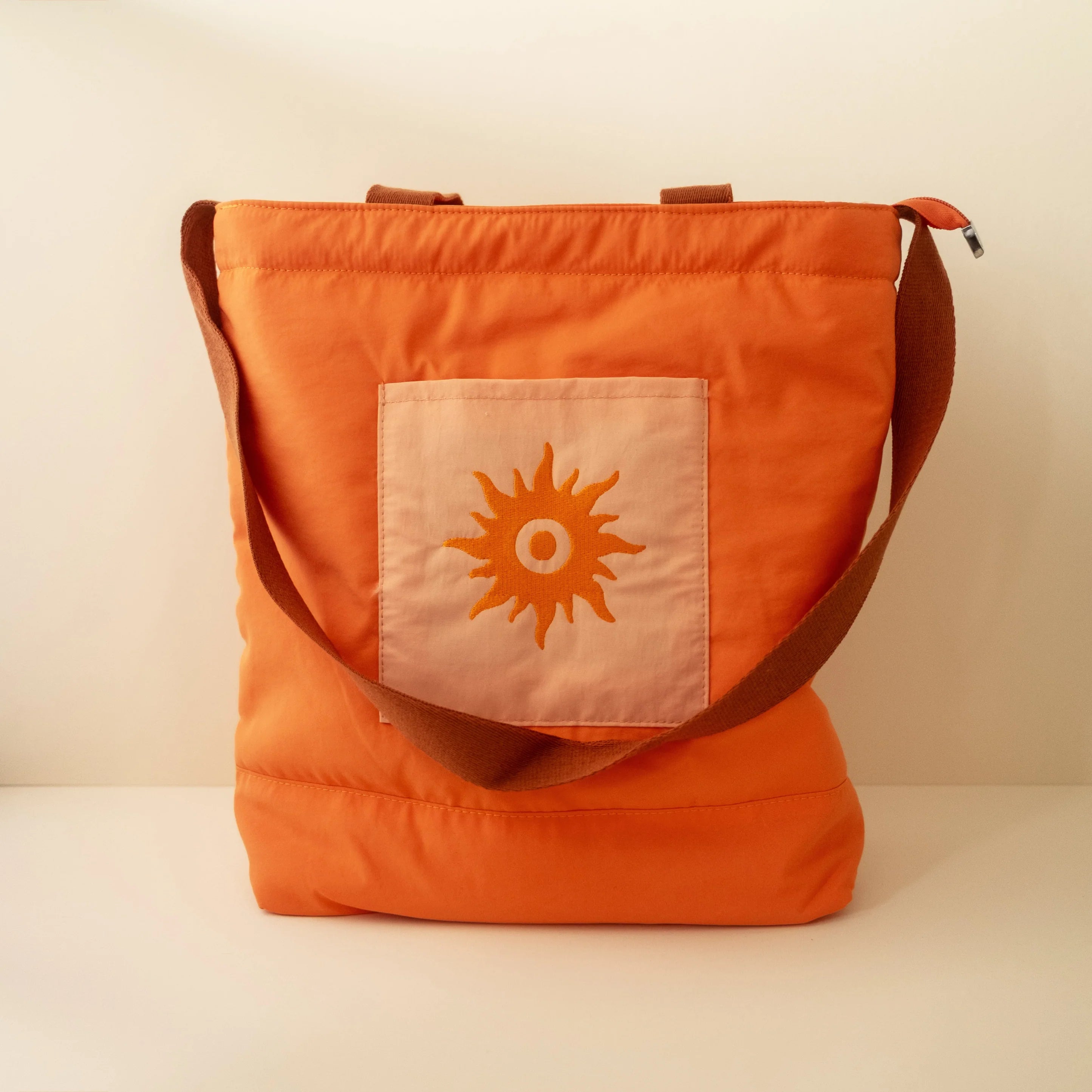 A burnt orange nylon beach tote with a sun pattern, featuring a top handle, a zipper closure, and an embroidered pocket on the front.