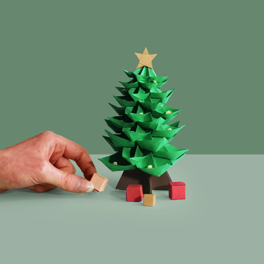 Hand placing a small box near a miniature origami Christmas tree on a green background