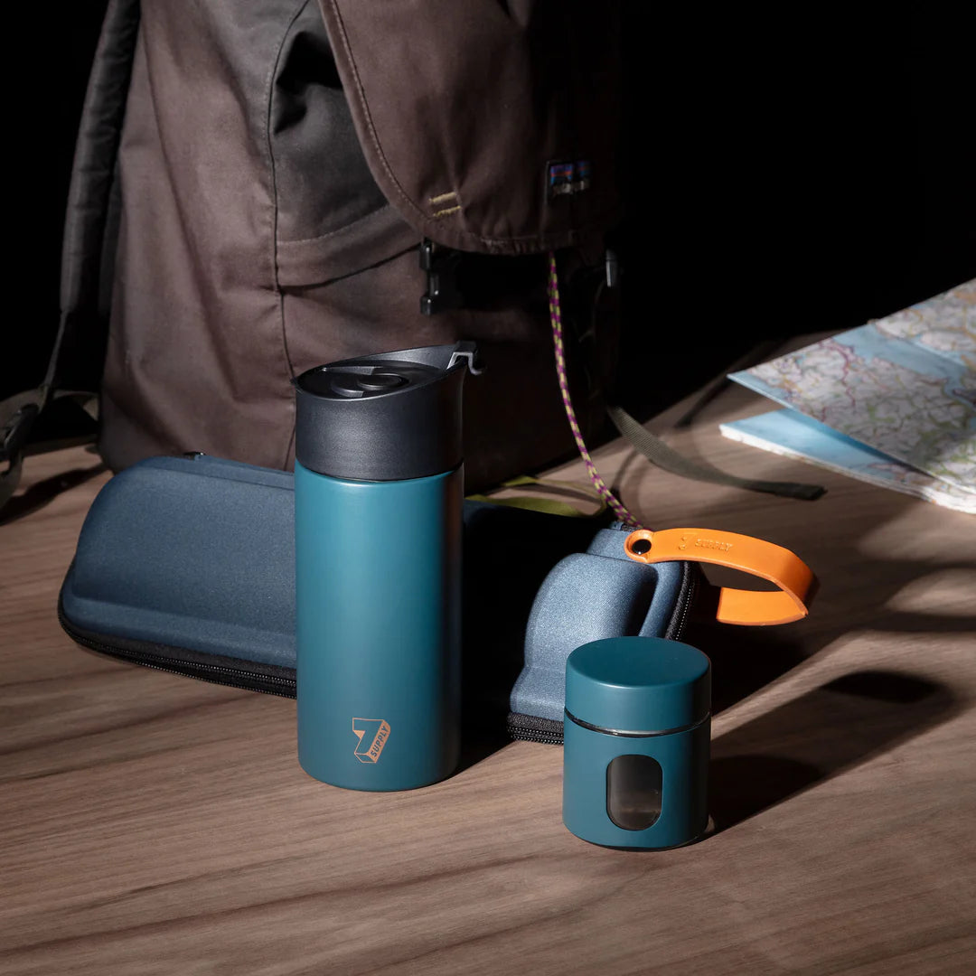 Portable French press coffee set with metal flask, color-coded jar for ground coffee, and matching travel case displayed on wooden surface and blurry background.