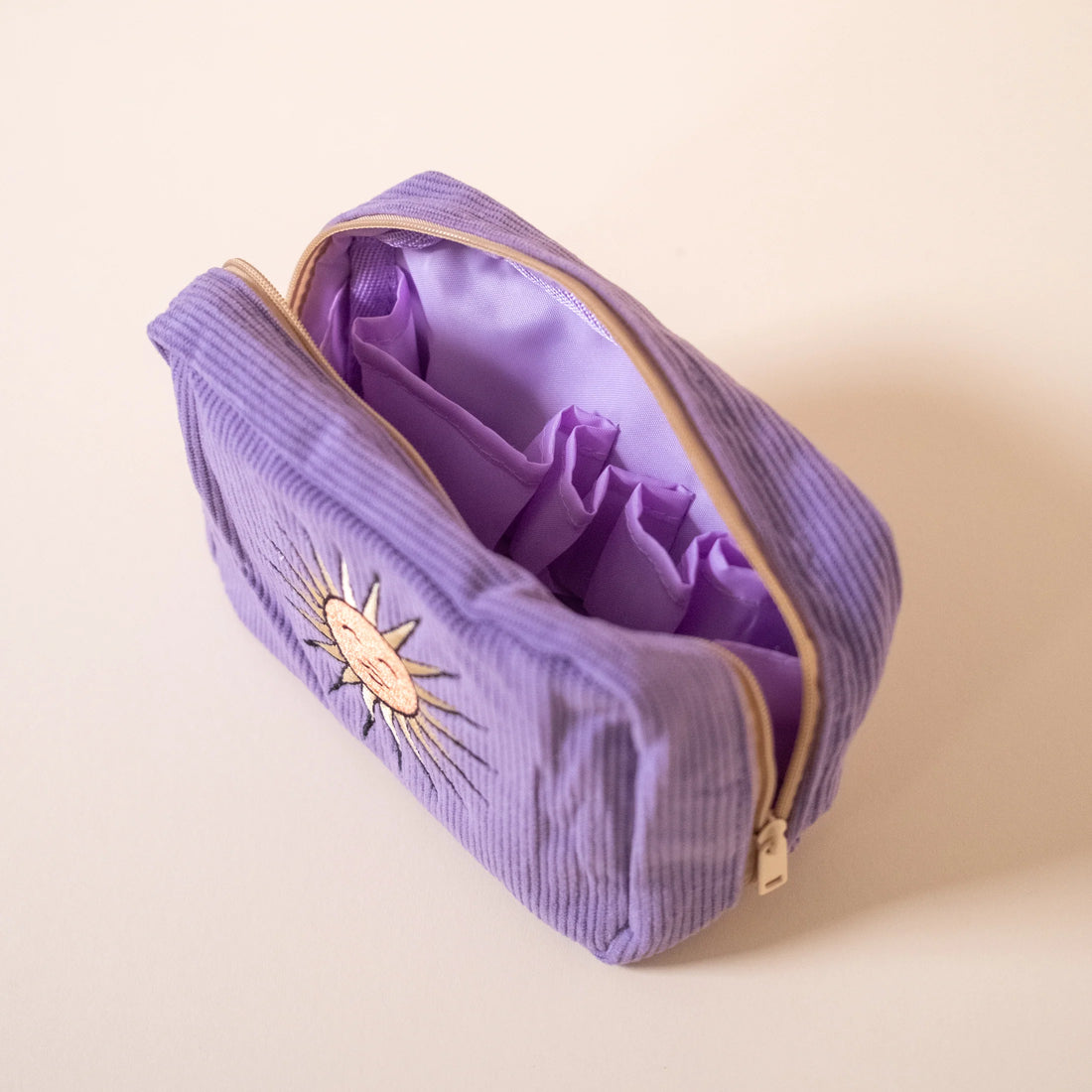 Open purple cosmetic bag with internal pockets and zipper, featuring sun embroidery on the front, displayed on a beige background