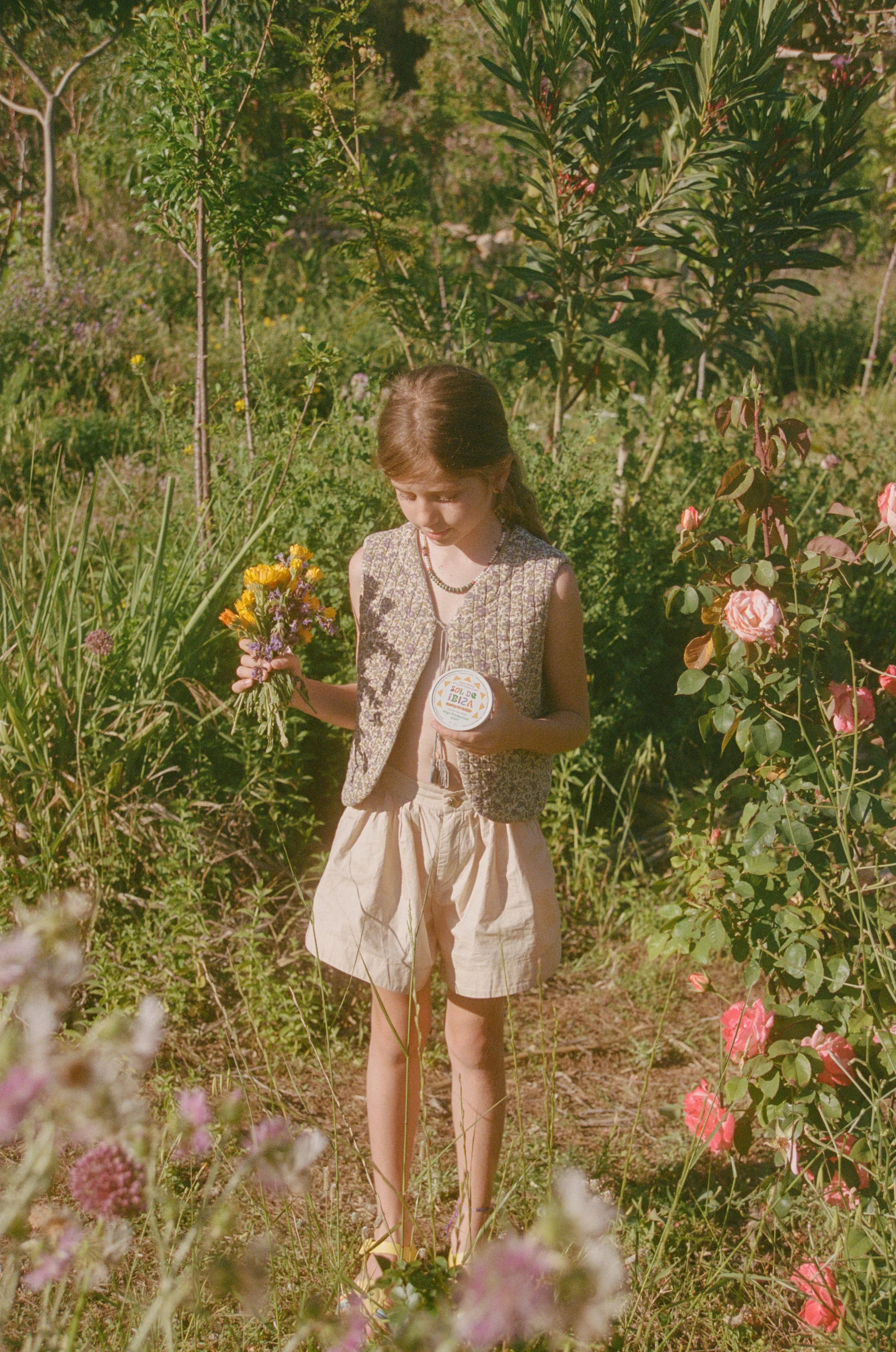 Young girl holding the Face & Body Plastic Free Tin Baby & Kids, and flowers in a garden with greenery and pink flowers around her.