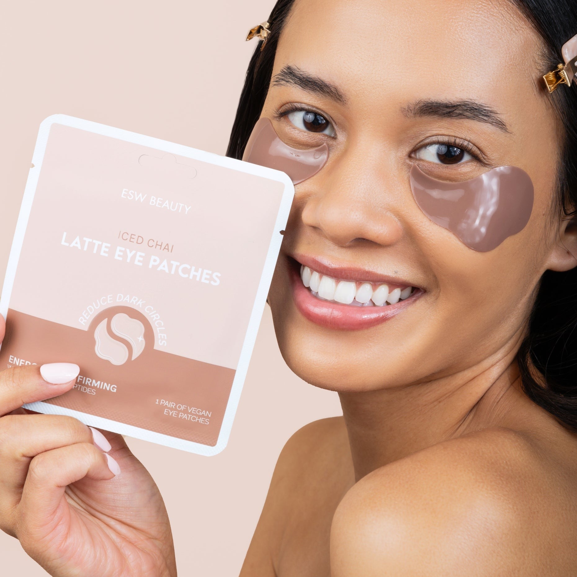 Woman wearing eye patches holding a package of eye patches against a beige background