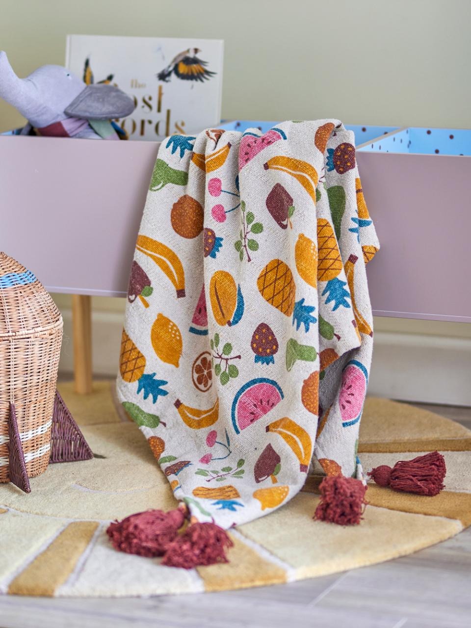 Colorful fruit-patterned blanket draped over a crib with a book and toys in the background.