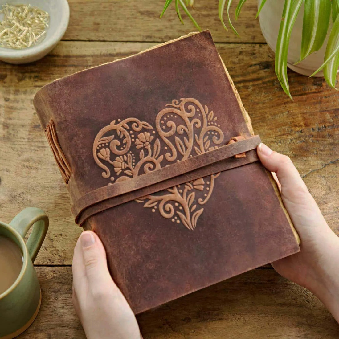 Two hands holding a Heart Buffalo Leather Wraparound Journal on a wooden surface, positioned near a green plant, showcasing the embossed heart design and wraparound leather cover.