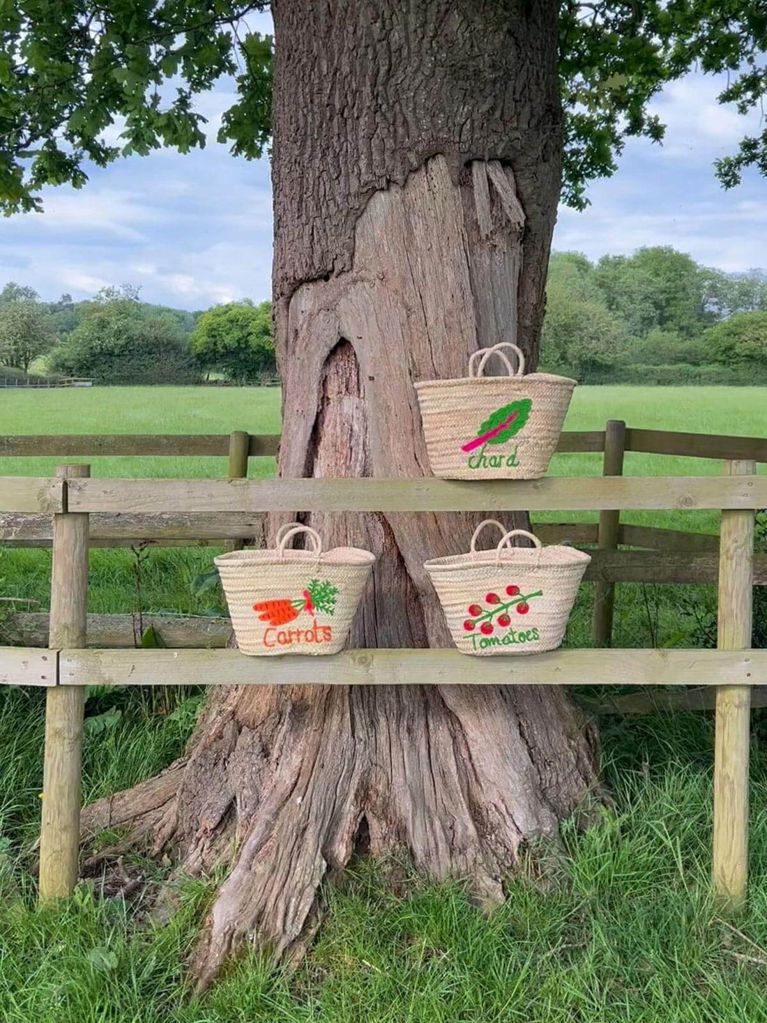 three handwoven palm leaf market baskets with embroidered designs of tomatoes, carrots, and chard in field scenes.