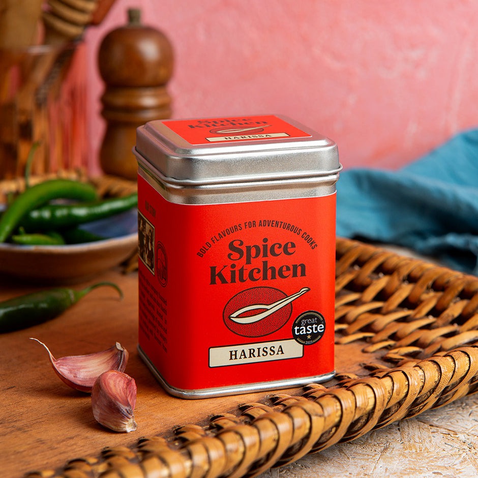 Red Spice Kitchen Harissa spice tin on a wooden surface with peppers and garlic in the background.