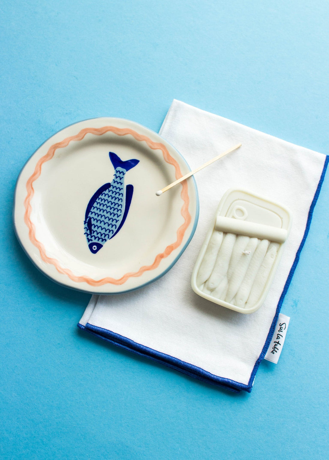 Tinned fish soy wax candle with blue fish decorative plate and matches, styled on a blue background
