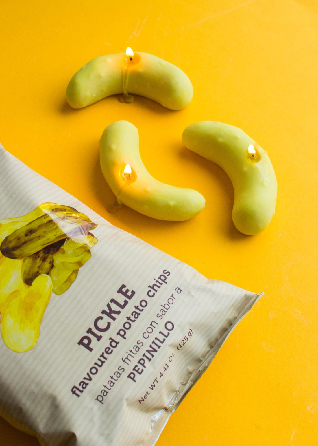 Green pickle-shaped soy wax candle with pickle crisp bag, styled on a yellow background