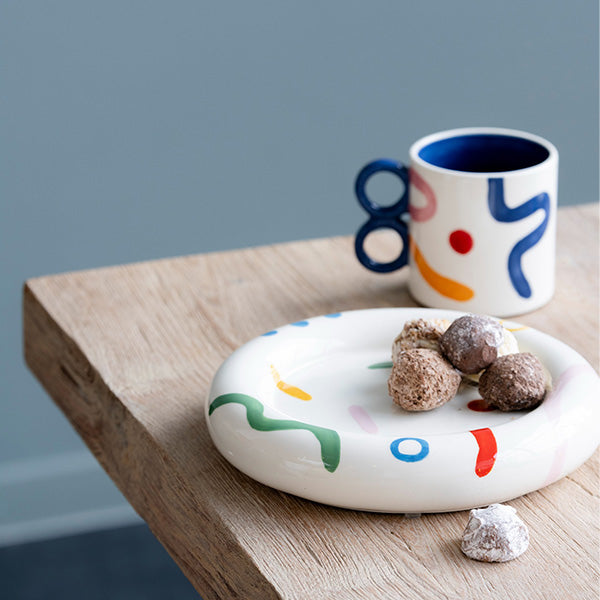 Sara Pintura Locura Mug with abstract modern hand-painted pattern in blue, red, pink, and orange with a matching plate next to it