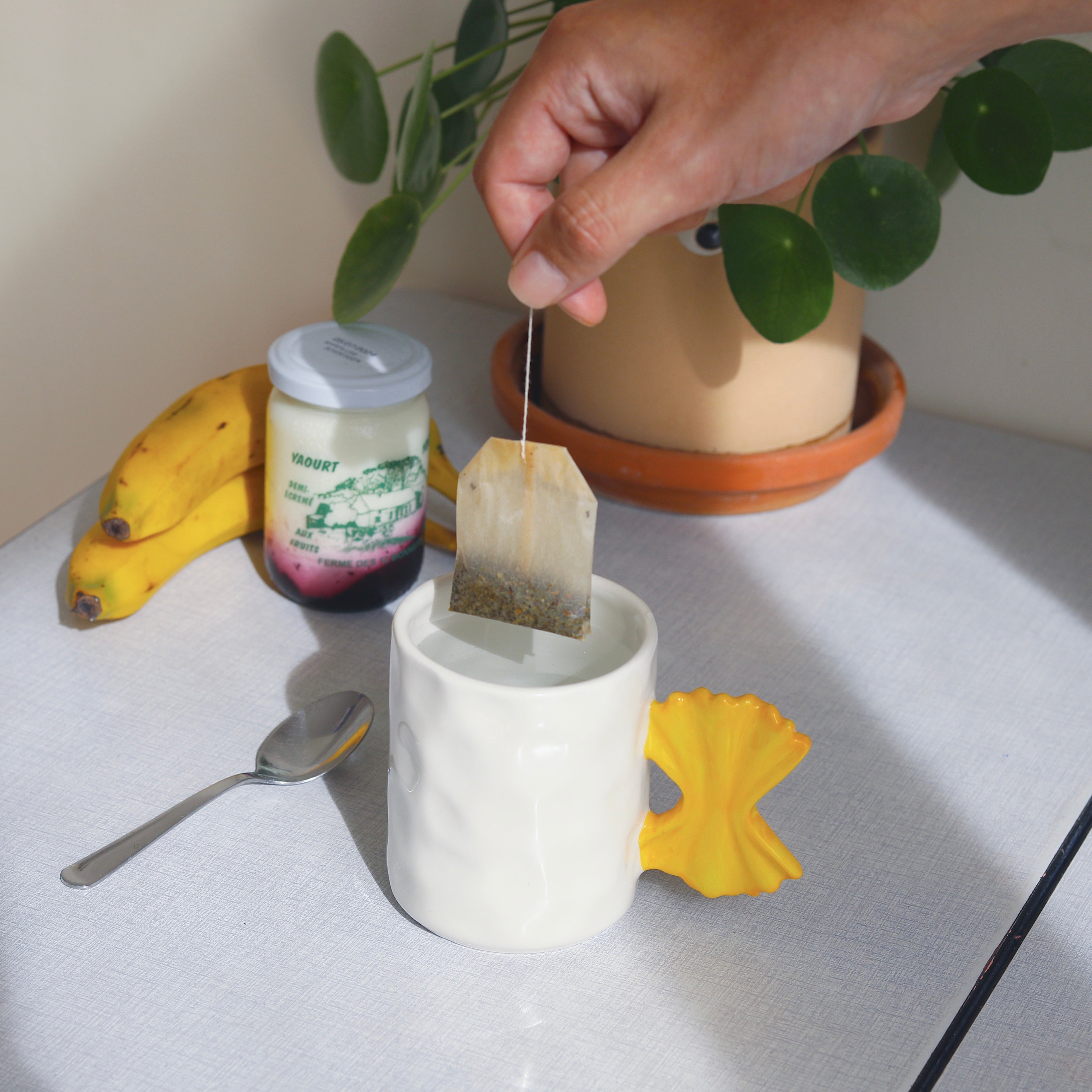 Tea bag being placed into a white mug with a yellow pasta-shaped handle, on a table with bananas, a jar, and a plant.
