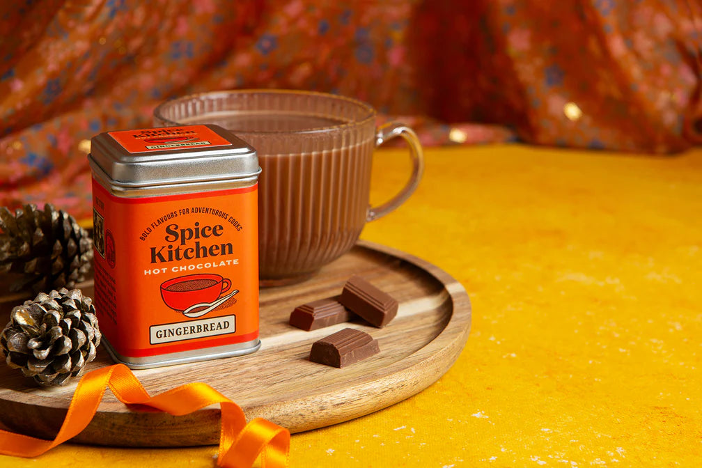 Spice Kitchen gingerbread hot chocolate with mug, and pieces of chocolates on a yellow  surface and orange background – vegan dairy-free hot chocolate.