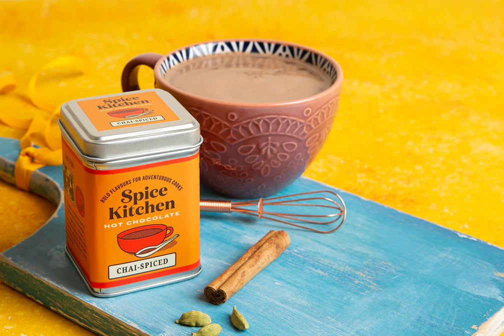 Spice Kitchen chai-spiced hot chocolate with mug, whisk, and cinnamon stick on blue surface and yellow background – vegan dairy-free hot chocolate.