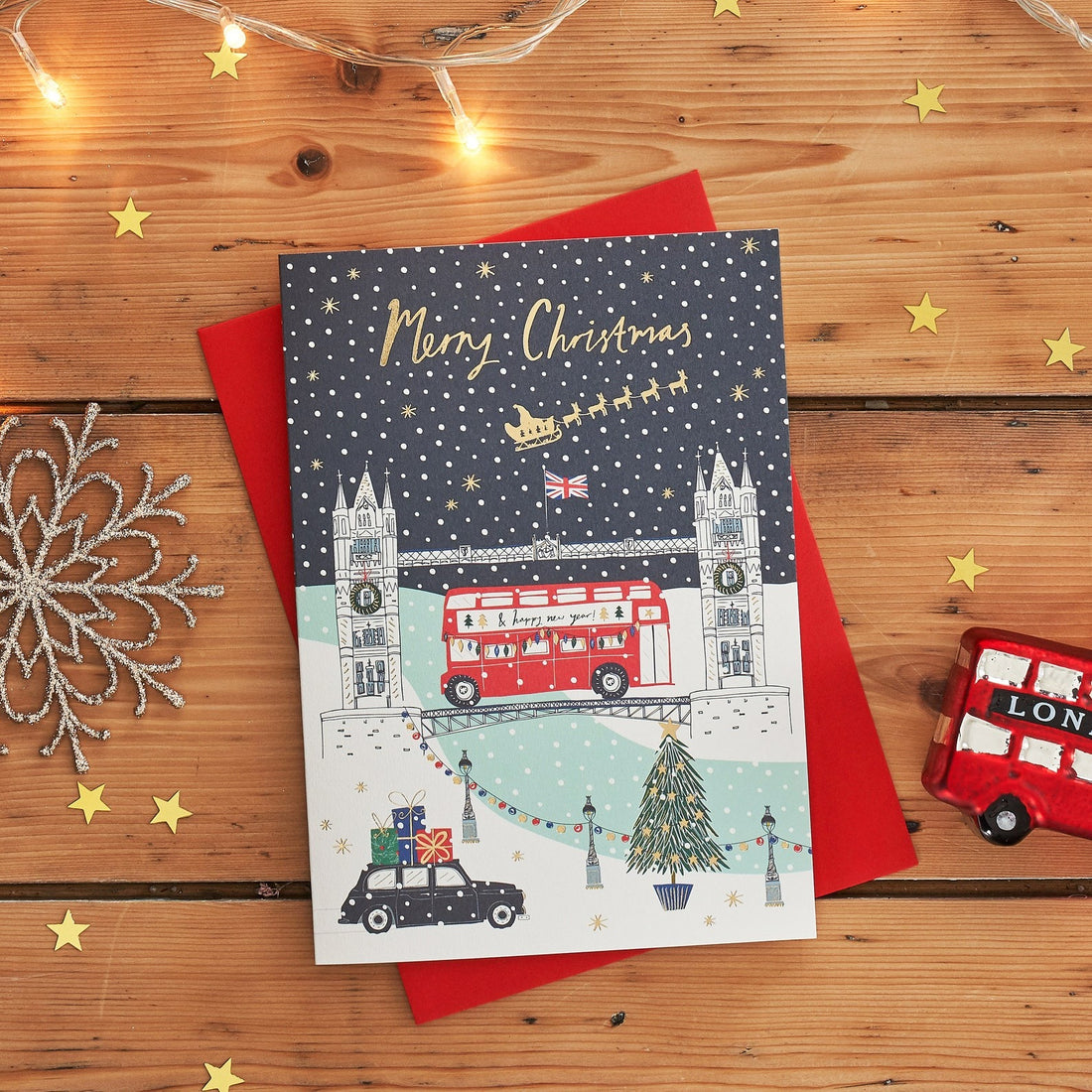 Gold Foiled Christmas Card featuring Tower Bridge with black cab in snowy London scene on a wooden surface