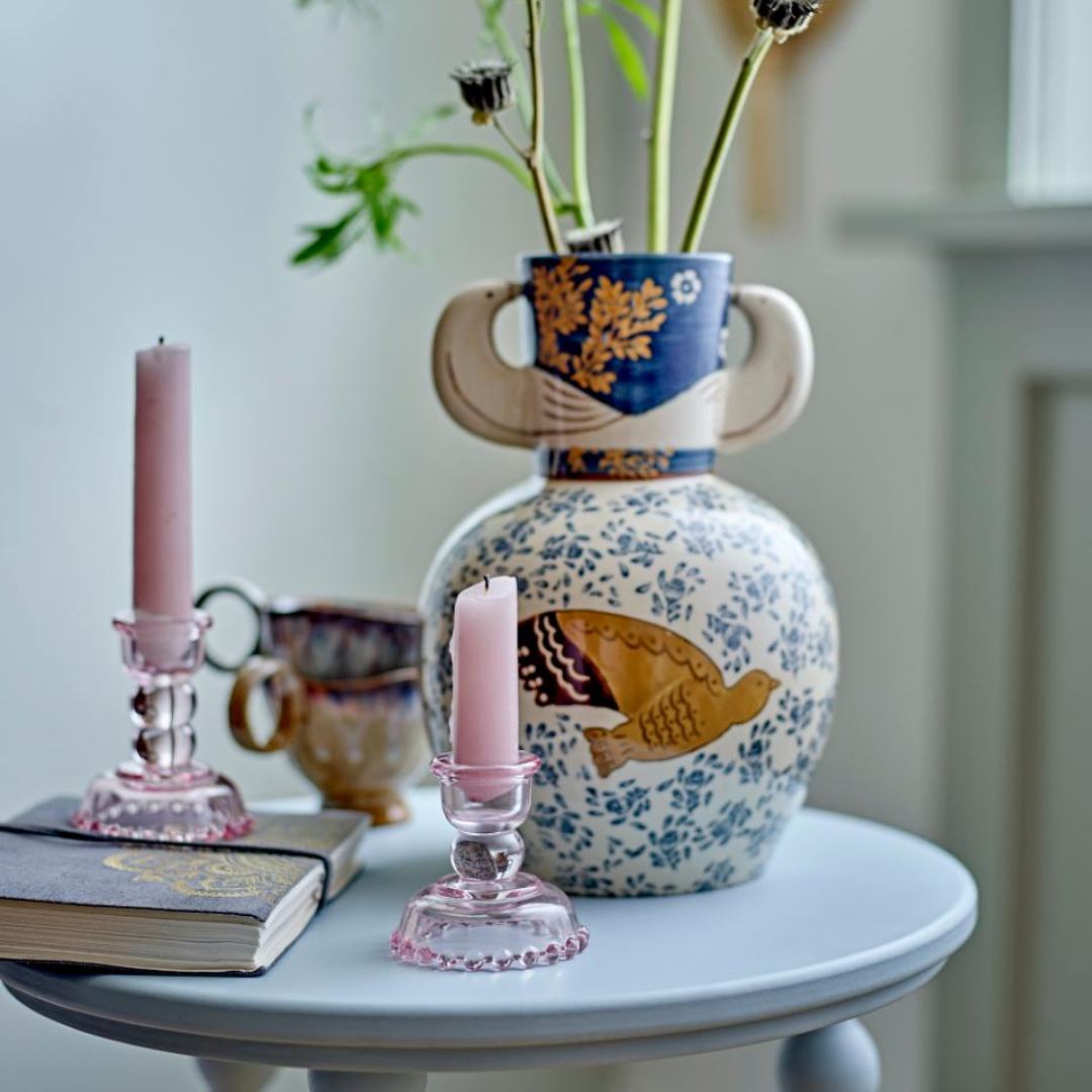 A blue and white ceramic vase with bird patterns, placed on a table alongside pink candlesticks, books, and other decorative items.