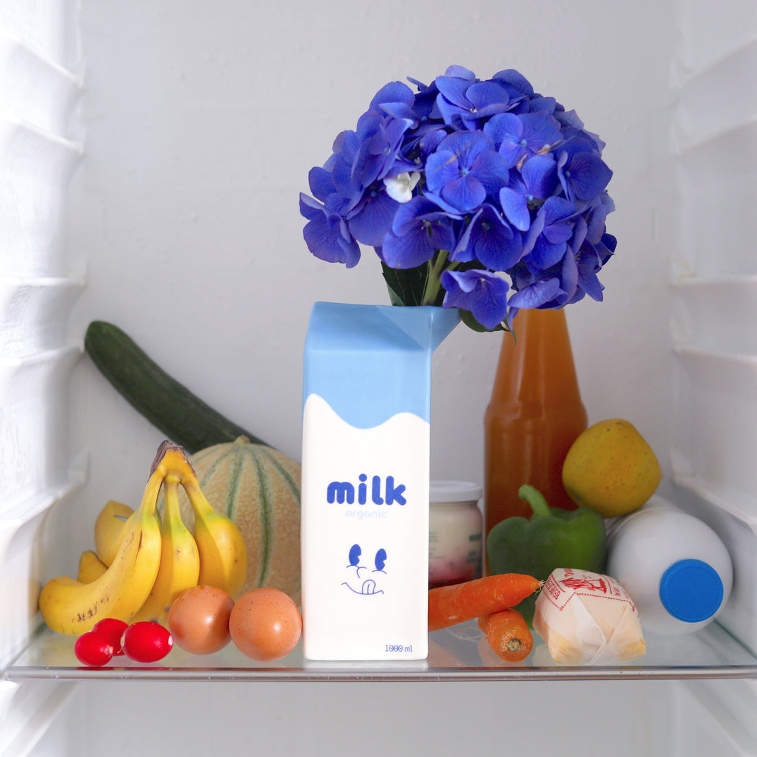 Fridge shelf with thed Glossy white ceramic milk carton vase, fruits, vegetables, and a vase of blue flowers.