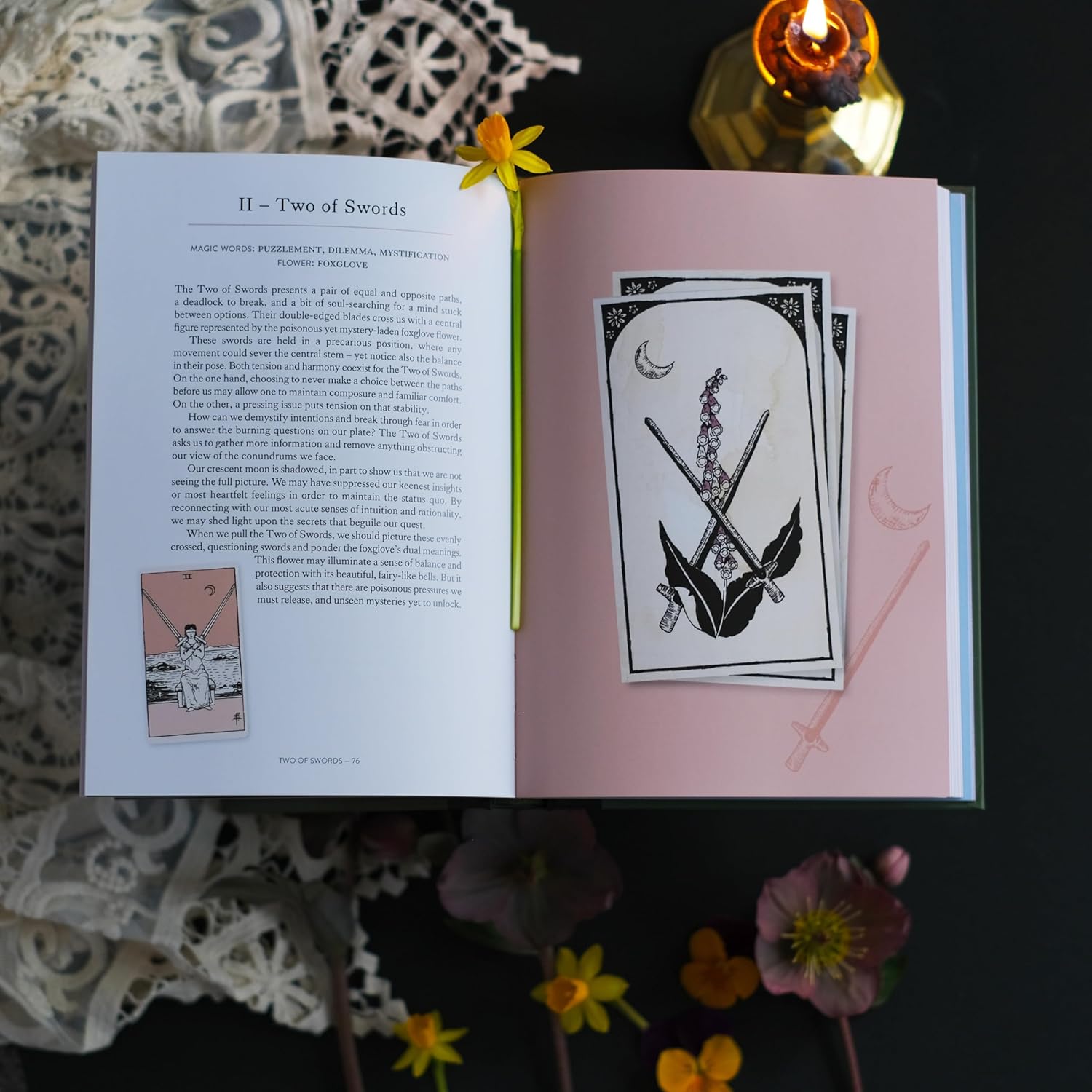Open book with tarot card illustration of two of swords and text on a dark surface with flowers.