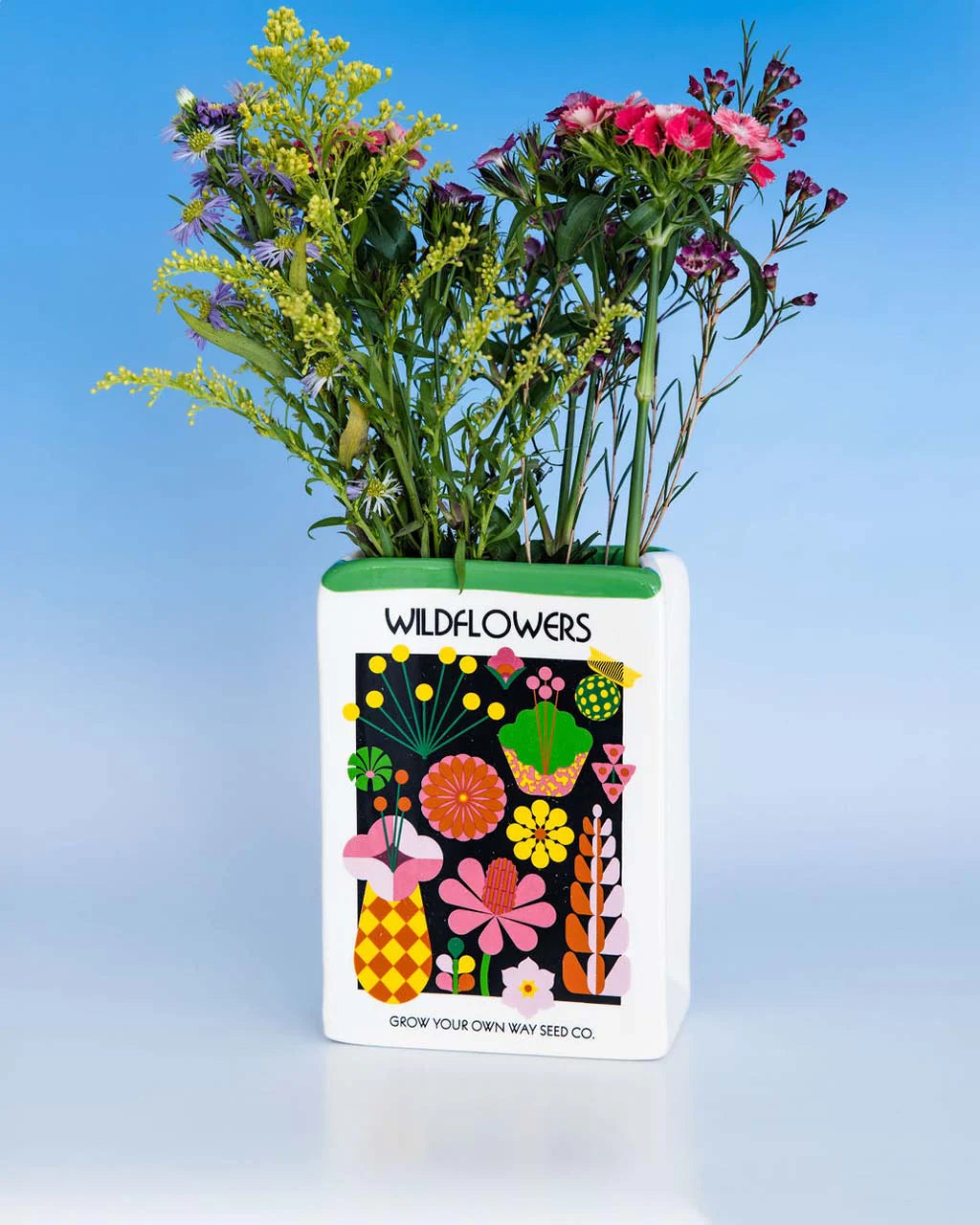 Decorative ceramic vase designed like a wildflower seed packet, filled with flowers, set against a blue background.