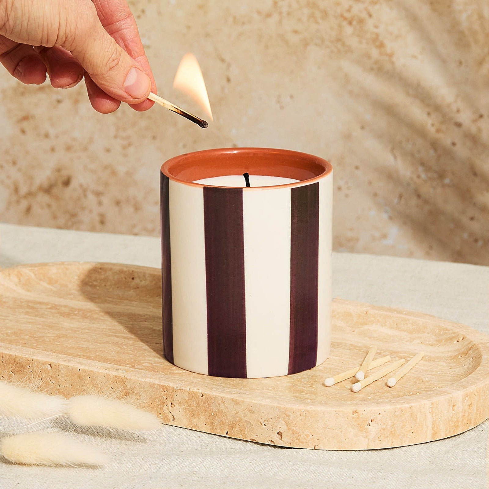 A person lighting the Marrakesh Chocolate Stripe Tomato Leaf Candle in a ceramic vessel