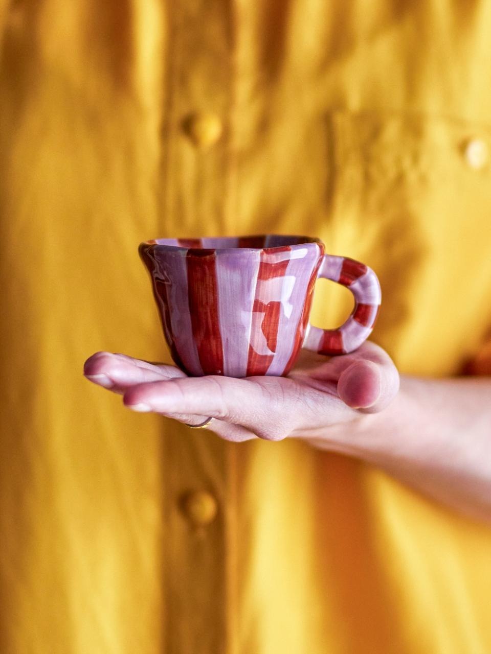 Hand holding a hand-painted small ceramic cup in pink featuring red stripes, against a yellow textured background
