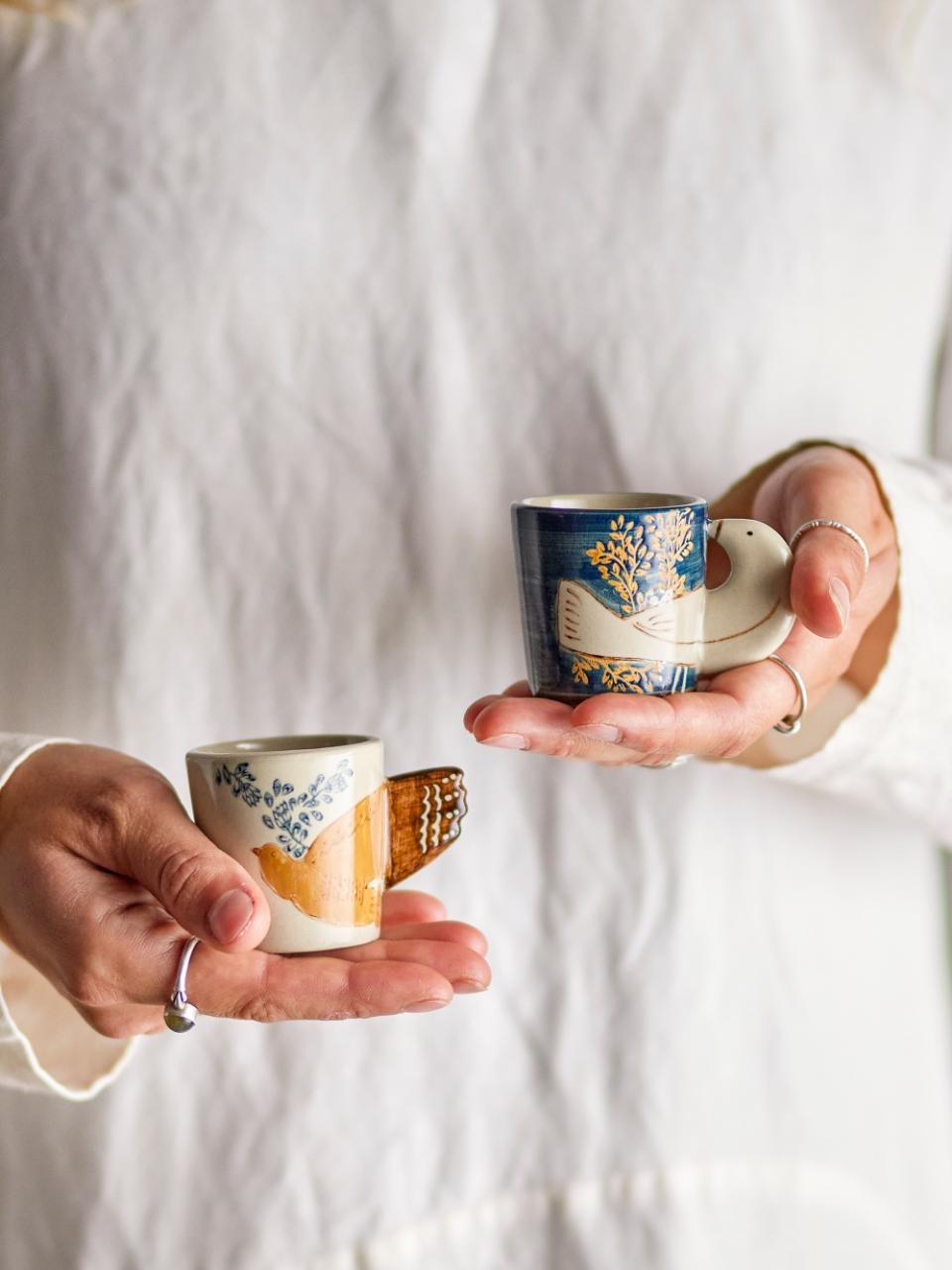 Two hands holding the two Hezha espresso cups, one in blue and one in nature color, each featuring a delicate bird motif with wings as handles. Hand-decorated, unique ceramic cups ideal for espresso or decorative display.