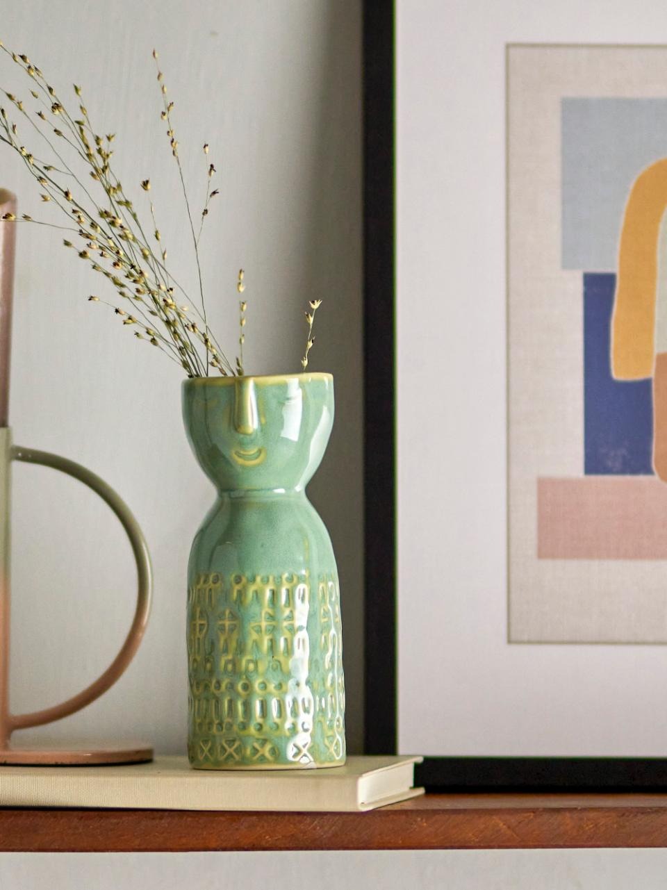 Green Stoneware Embla Vase with dried flowers, displayed on a white book with a blurred painting in the background – minimalist home décor setting