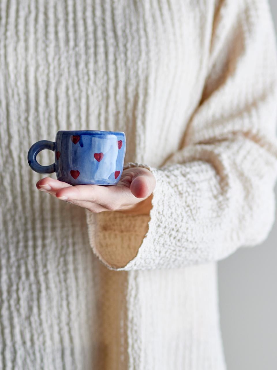Person holding a small blue mug with red hearts against a neutral background