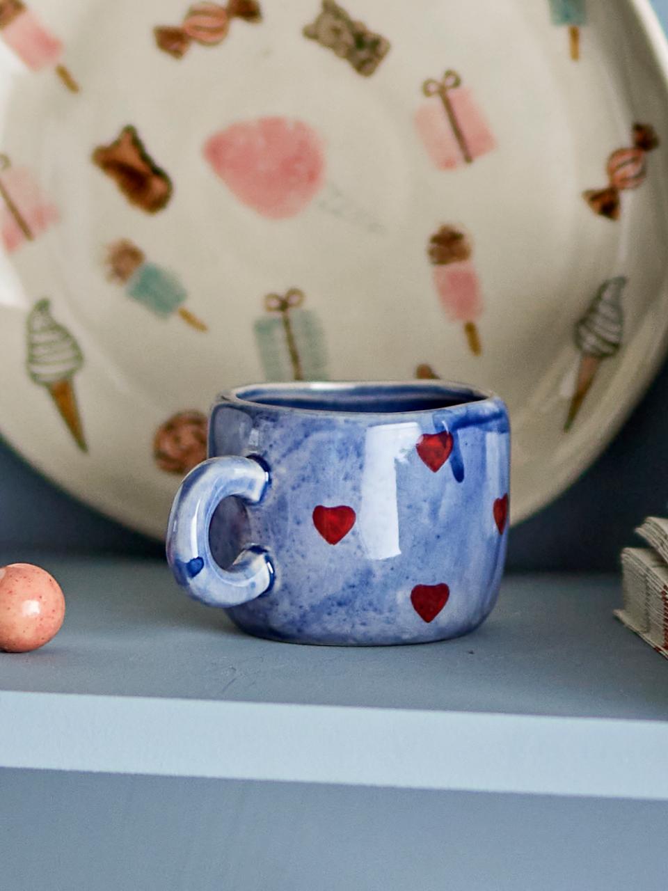 Blue ceramic mug with red heart designs on a gray surface, with a patterned plate in the background.