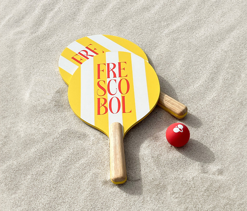 Two yellow and red striped Frescobol beach rackets with wooden handles and a red ball, plus a striped cloth bag on a beach sand.