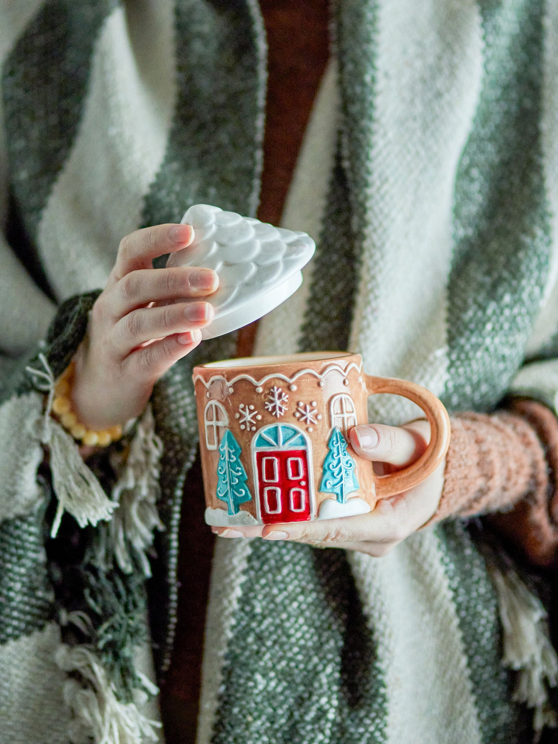 Person holding the Stoneware Hand-painted Ginnie Mug in brown with a charming gingerbread house design and roof-shaped lid, perfect for coffee, tea, or hot chocolate.