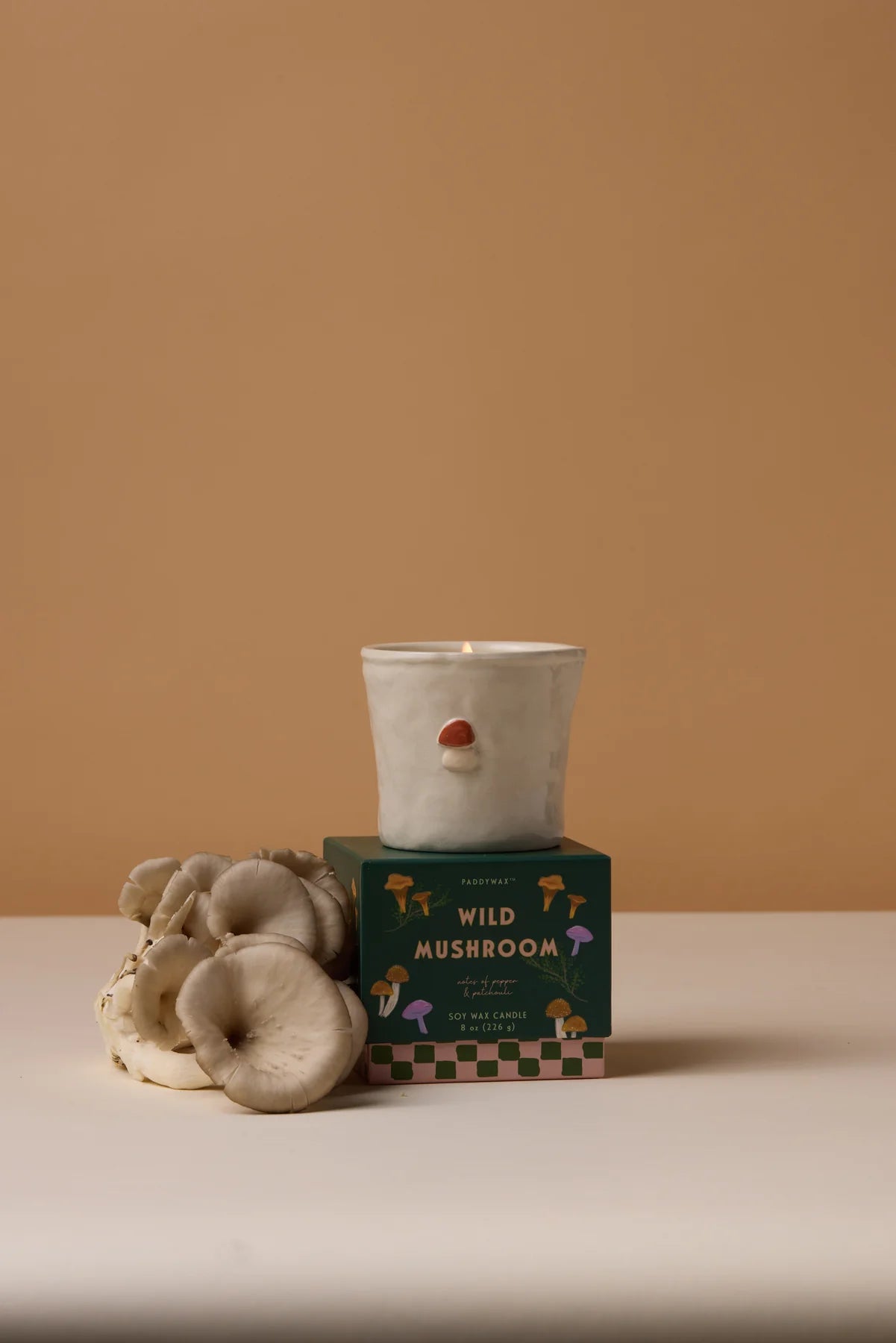 Candle in a white holder with handpainted mushroom on top, placed on a green box labeled &