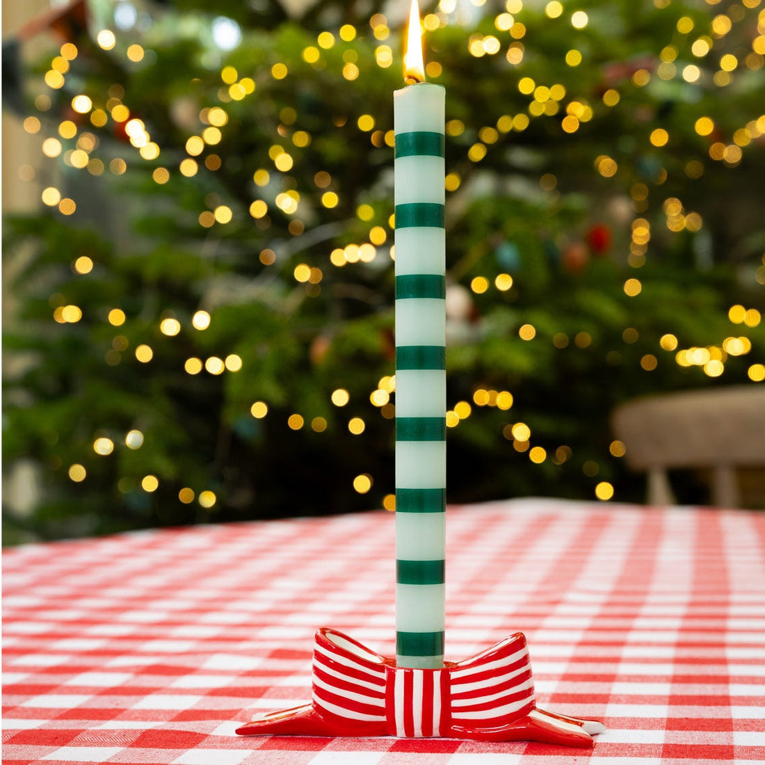 Green and white striped candle on a red and white striped bow candle holder with a blurred Christmas tree in the background.