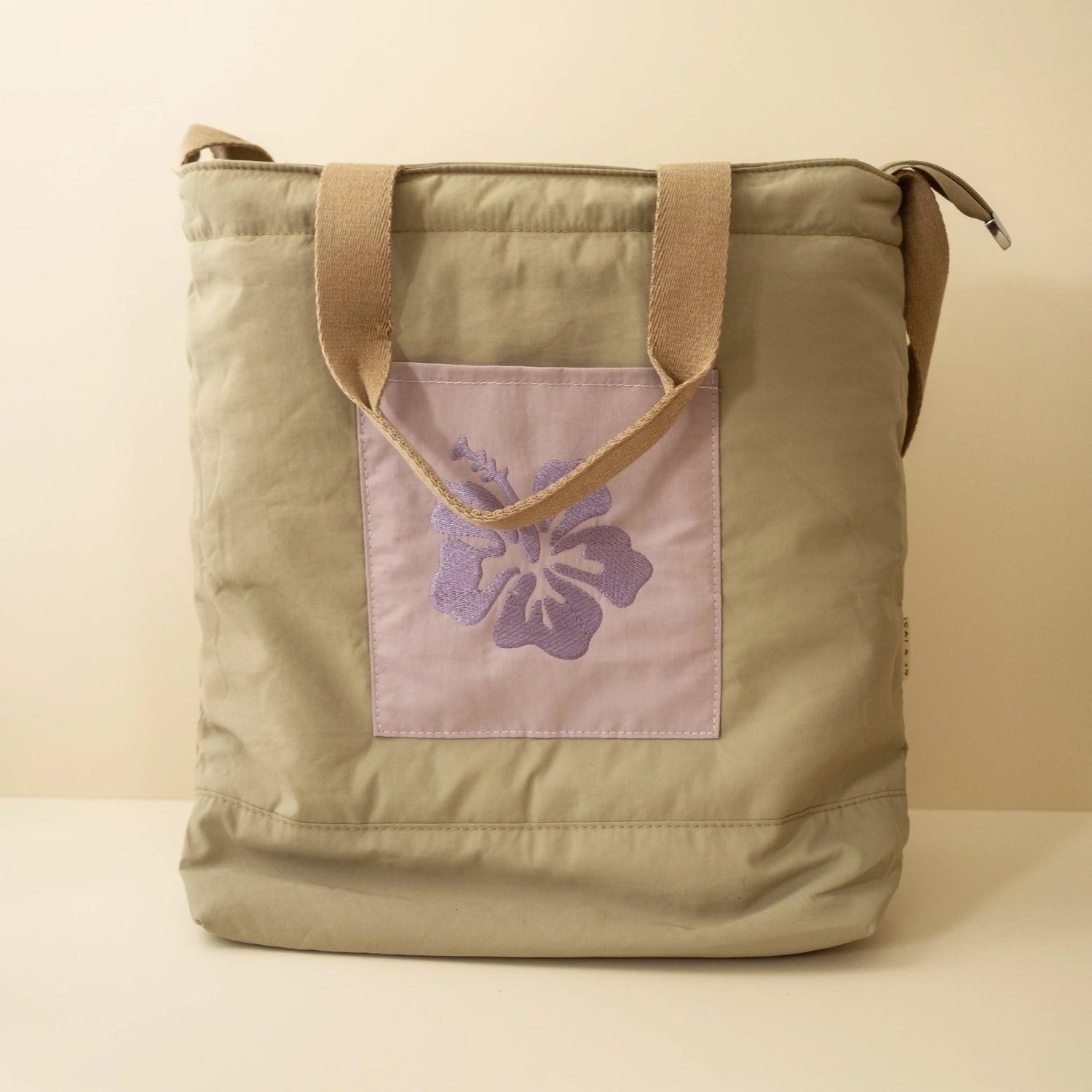Olive green nylon beach tote with a purple floral embroidery on the front pocket and two long handles.
