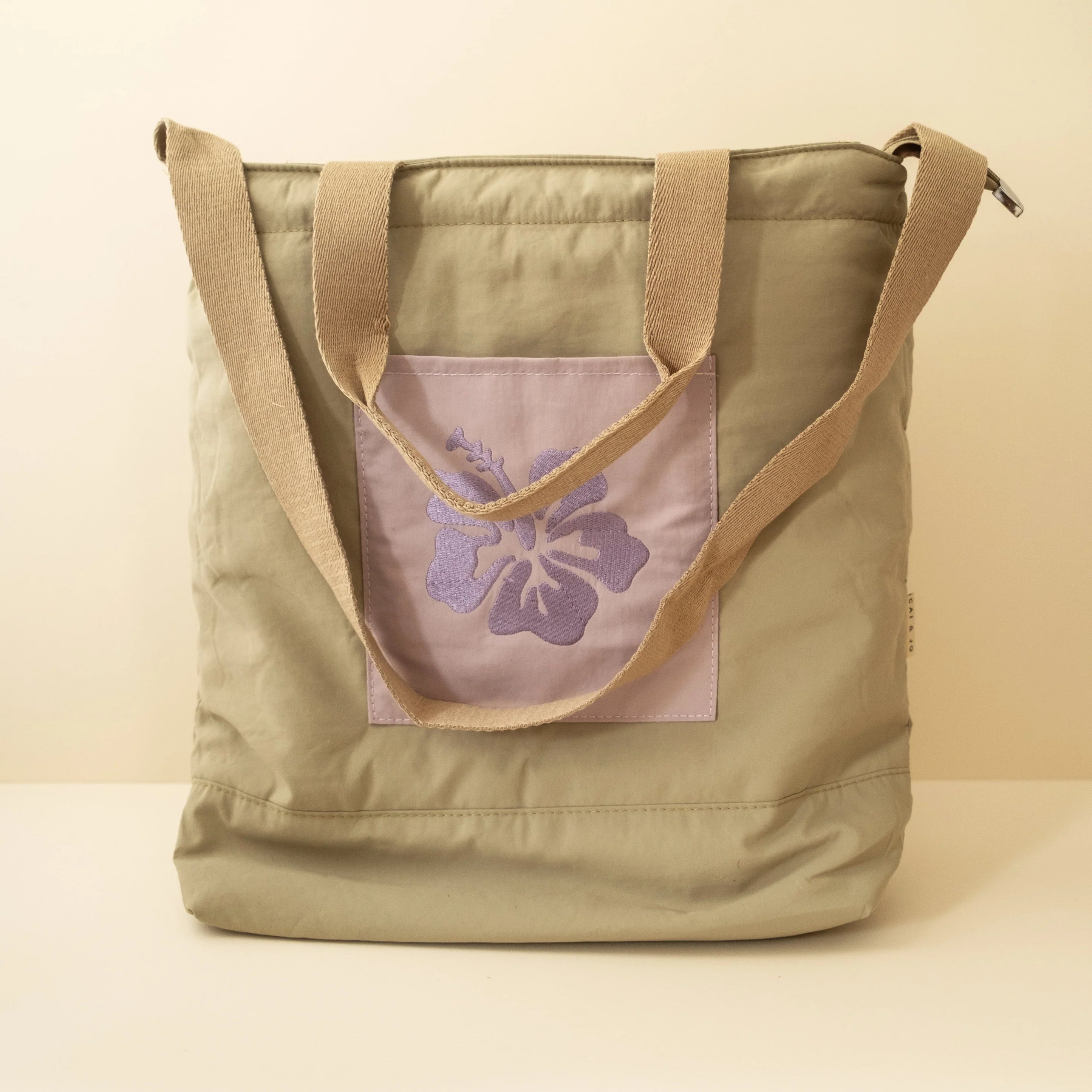 Olive tote bag with two brown handles and a purple embrodery flower design on the front pocket on a beige background