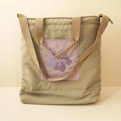 Olive tote bag with two brown handles and a purple embrodery flower design on the front pocket on a beige background