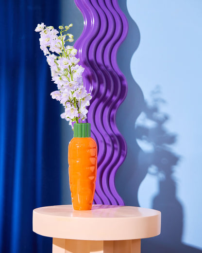Carrot-shaped vase with flowers on a round table against a blue and purple gradient background