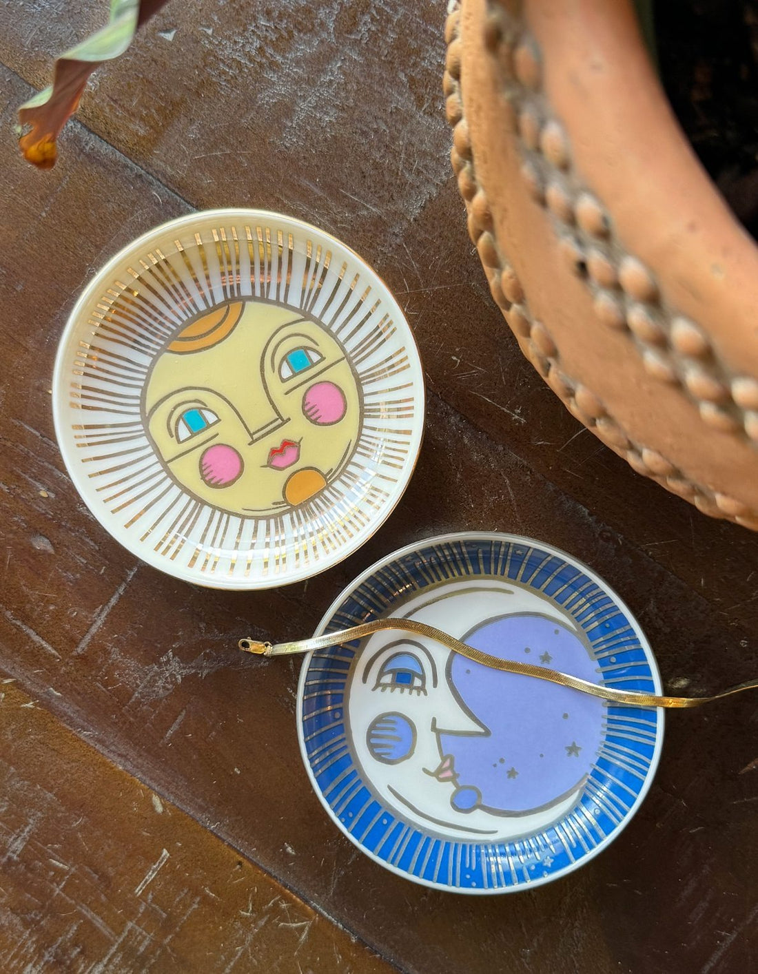 Moon and Sun porcelain trinket dishes displayed on a wooden surface, with a gold bracelet resting on top of the Moon dish, showcasing hand-painted designs and luxurious metallic detailing.