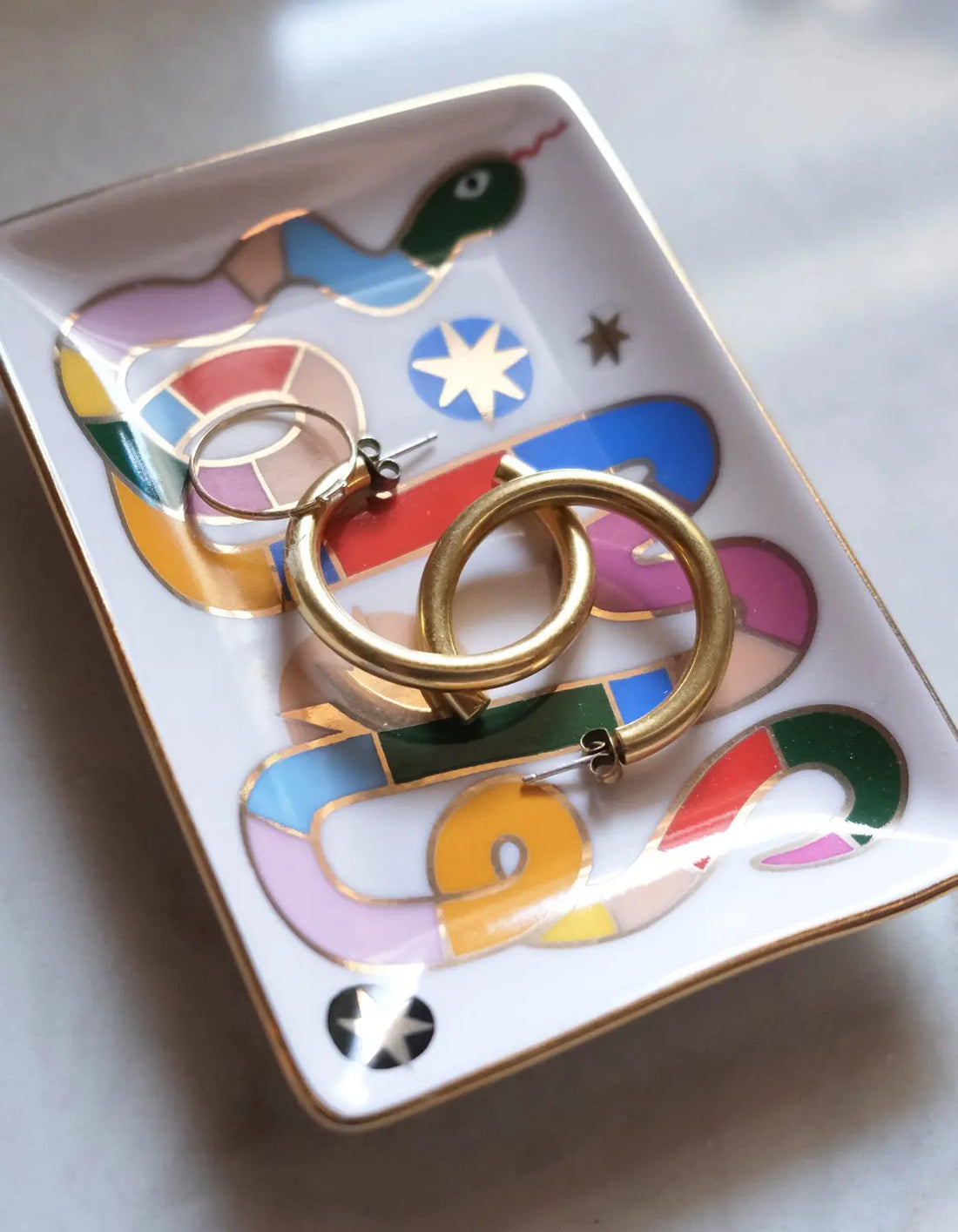 Compact ceramic trinket dish with hand-painted rainbow snake design and 10k gold detailing, holding earrings and small jewelry, ideal for keys or other essentials, presented in an eco-friendly gift box.